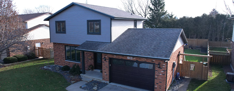Beautifully Renovated Home In Chatham Ontario, featuring Stonecrest Grey vinyl siding, dark engineered wood trims, black window capping's, soffit, fascia and eavestrough, and a mahogany woodgrain front door with wrought iron window accents and Gemstone Soffit Lighting.